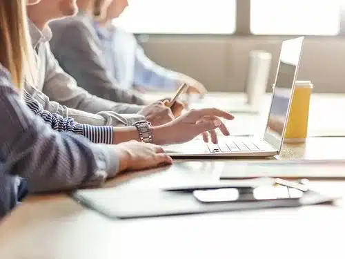 Mehrere Personen sitzen an einem Tisch, arbeiten an Laptops und Tablets und machen Notizen; Fokus liegt auf den Händen und Geräten.