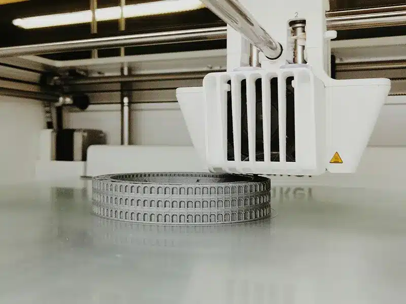 Ein 3D-Drucker erstellt ein rundes, graues Bauteil mit regelmäßigen, bogenförmigen Vertiefungen auf einer glatten Druckplatte.